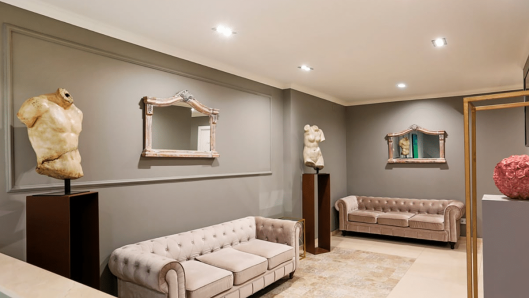 Sala de espera del interior de Monarka Clinic con paredes grises paneladas, focos empotrados en el techo, sofás beige capitoné, esculturas clásicas de torsos sobre pedestales, espejos decorativos y una alfombra en tonos neutros.