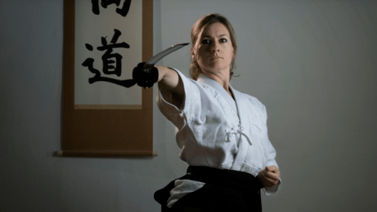 La Dra. Mireia Illueca Moreno practicando artes marciales con uniforme blanco y cinturón negro, realizando una técnica con espada, con un pergamino con caligrafía japonesa al fondo.
